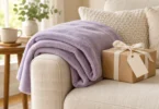 A plain lavender fleece throw draped over a cream sofa beside a ribbon-tied gift box with a blank tag