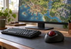 A black trackball with a dark red ball sits beside a compact keyboard on a wooden desk, with a colorful landscape map on the monitor.