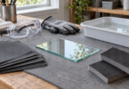 A loose rectangular glass sample rests on a gray work mat beside abrasive sheets, foam hand pads, protective goggles, gloves and a water tray.