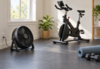A black floor fan beside an exercise bike, rolled mat and dumbbells in a bright home workout room.