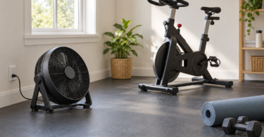 A black floor fan beside an exercise bike, rolled mat and dumbbells in a bright home workout room.