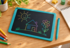 A generic teal doodle tablet shows colorful line drawings of a sun, house and flower beside a matching stylus and colored pencils on a wooden table.