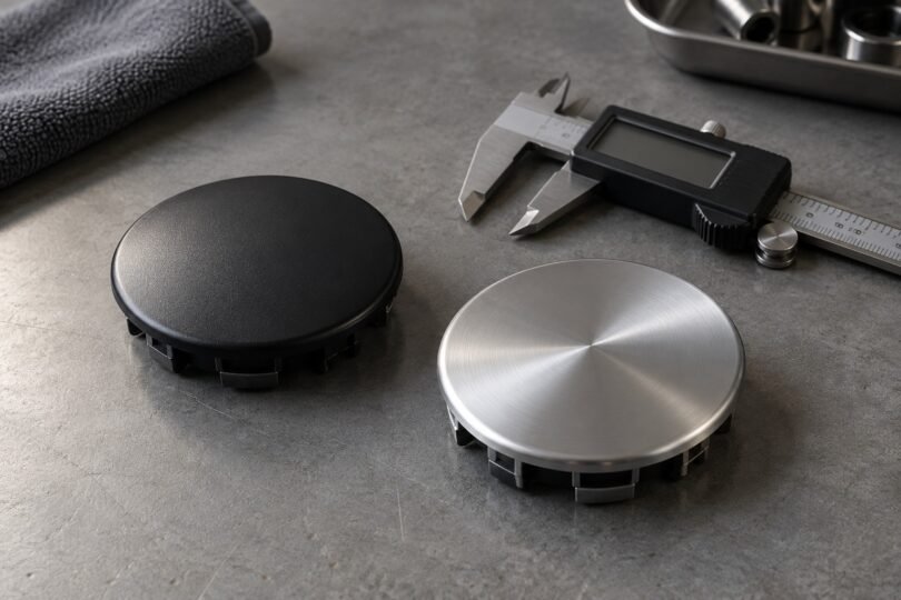 Unbranded black and silver round wheel center caps beside a digital caliper on a gray workbench