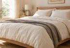 A bed with unbranded cream bedding, matching pillow shams and a gray throw in a sunlit neutral bedroom