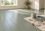 Illustration of a sage-gray painted plank floor beside an open window, with a closed plain paint can, roller, brush, gloves and safety glasses on a drop cloth.