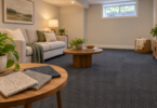 Three neutral carpet swatches on a wooden table in a sitting room with a sofa, high window and dark square-patterned carpet floor.