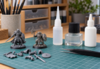 Generic miniature figures, loose model parts and three closed adhesive bottles on a cutting mat beside tweezers