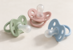 Three unbranded pacifiers in sage green, dusty pink and pale blue on a clean cream surface.