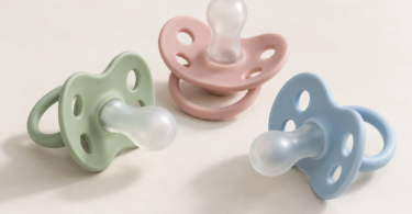 Three unbranded pacifiers in sage green, dusty pink and pale blue on a clean cream surface.