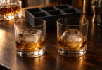 Two lowball glasses with amber drinks, one holding a large ice cube and the other an ice sphere, beside a dark ice tray.