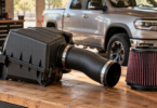 Black airbox, intake tube and pleated filter on a workbench with a silver pickup behind them