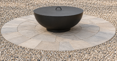 Covered black metal fire bowl on a circular paved pad surrounded by beige and gray landscaping gravel