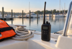 A dark handheld radio with a long antenna stands beside coiled dock line and an orange life jacket on a boat at a calm marina.