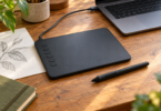 A black screenless drawing tablet and stylus on a wooden desk beside a laptop and a pencil sketch of leaves.