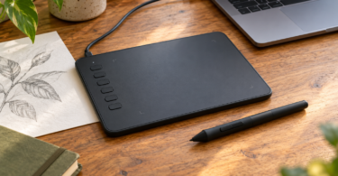 A black screenless drawing tablet and stylus on a wooden desk beside a laptop and a pencil sketch of leaves.