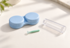 Illustration of a closed blue contact lens case, a small green cup-tipped handling tool and a clear protective tube on a light surface