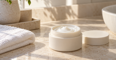 An open unbranded jar of white face cream with its lid and a folded towel on a sunlit stone bathroom counter