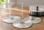 Three generic blank compact discs beside an open clear jewel case and a spindle on a wooden table