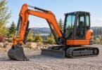Unbranded mini excavator parked on gravel with its bucket grounded and thumb folded against the stick