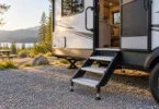 Three metal entry steps beside the open doorway of a white travel trailer at a wooded campsite