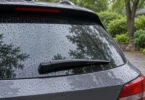 Black rear wiper resting on rain-covered hatch glass beside a leafy driveway