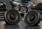 Two generic round speakers on a workbench with a parked touring motorcycle and helmet in the background.