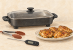 Conceptual illustration of a covered unbranded electric skillet, a plate of golden chicken, silicone-tipped tongs and a food thermometer on a cream counter.