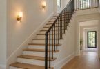 Two brass wall sconces with pale shades illuminate a cream wall beside an oak staircase with white risers and a black metal railing.