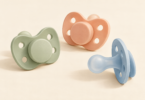 Conceptual illustration of three unbranded infant pacifiers with vented shields in sage, peach and blue on a cream surface.