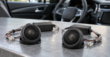 Two generic black automotive tweeters and inline crossovers on a workbench beside a car interior