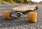 Close-up of amber wheels and a metal truck beneath a wooden longboard resting on coarse asphalt.