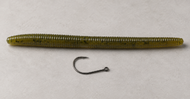 One unbranded dark fishing hook beside an olive-green soft-plastic worm on a neutral studio surface