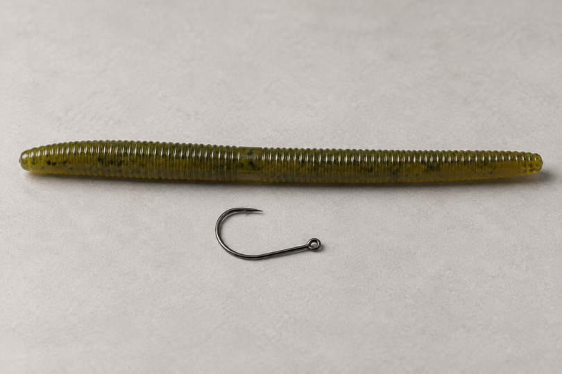 One unbranded dark fishing hook beside an olive-green soft-plastic worm on a neutral studio surface