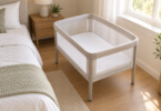 Empty mesh-sided bassinet with an upright enclosed rim and flat white fitted sheet, standing separately beside an adult bed