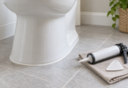 White toilet base on gray tile with an unbranded caulk gun and finishing tool on a folded cloth