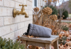 Unconnected brass outdoor faucet on a pale brick wall above a gray insulated pouch resting on a wooden stool