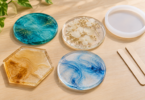 Four translucent resin coasters in teal, clear floral, amber and pale blue designs beside an empty white mold and two wooden mixing sticks