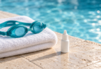 Teal swim goggles on a folded white towel beside a capped, unbranded drop bottle at a swimming pool.
