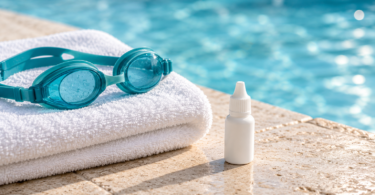 Teal swim goggles on a folded white towel beside a capped, unbranded drop bottle at a swimming pool.