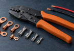 Generic orange-handled crimping tool beside copper ring lugs, silver contacts and disconnected red and black wire pieces on a dark workbench.