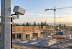 A plain outdoor camera housing secured to a steel post overlooking unfinished timber framing and a distant crane.