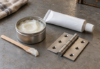 White grease in an open tin beside an unbranded tube, metal hinge and wooden applicator on a workbench.