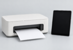 Unbranded white desktop printer with a blank sheet beside a black tablet on a pale gray background