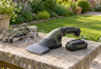 Covered cordless grass shear, detached battery, gloves and safety glasses on a stone bench beside a lawn