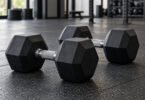 Illustration of two black hex dumbbells with silver handles on a rubber gym floor