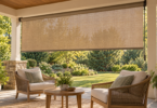 A tan woven roller shade hangs above two cushioned chairs and a round wooden table on a covered patio facing a leafy garden.
