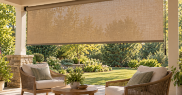 A tan woven roller shade hangs above two cushioned chairs and a round wooden table on a covered patio facing a leafy garden.
