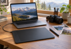 An unbranded screenless pen tablet and stylus sit on a wooden desk beside a laptop displaying a mountain photo, a camera and photographic prints.