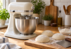 Unbranded cream stand mixer with a stainless bowl beside three pizza dough balls, flour and a bench scraper on a wooden worktop.