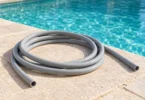 A smooth gray hose coiled on a pale stone deck beside turquoise swimming-pool water